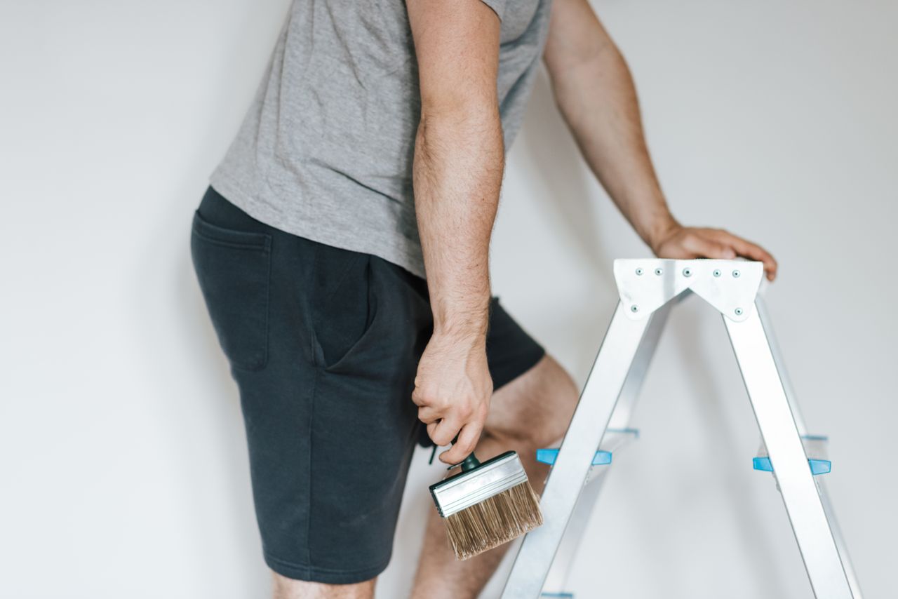 Side view of cropped anonymous male decorator in casual clothes getting ready to paint the walls of a bright room while standing on a stepladder